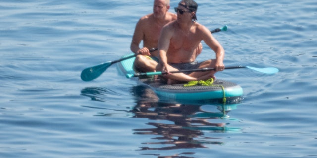 Woody Harrelson paddled in the back of the long board wearing neon blue and green striped shorts.