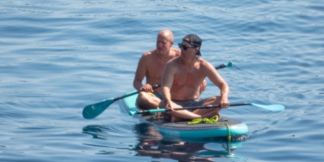 Woody Harrelson paddled in the back of the long board wearing neon blue and green striped shorts.