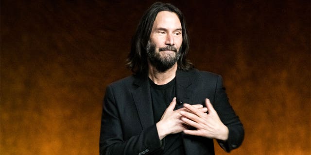 Keanu Reeves in a black blazer and t-shirt puts his hands to his chest (near his heart) on stage