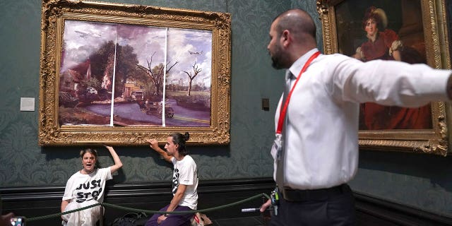 A security guard looks at protesters who glued their hands to the frame of John Constable's The Hay Wain, inside the National Gallery, London, Monday July 4, 2022. Police say two climate change protesters have been arrested after they glued themselves to the frame of a famous John Constable painting hanging in Britain’s National Gallery. The two, from the protest group "Just Stop Oil," stepped over a rope barrier and covered "The Hay Wain" on Monday with large sheets of paper depicting "an apocalyptic vision of the future" of the landscape. 