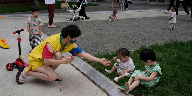 A woman wearing a mask tries to take photos for children as they play on the lawn, Wednesday, July 6, 2022, in Beijing. 