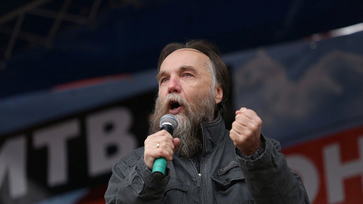 Putin's brain, Alexander Dugin, holds a microphone while wearing a brown coat