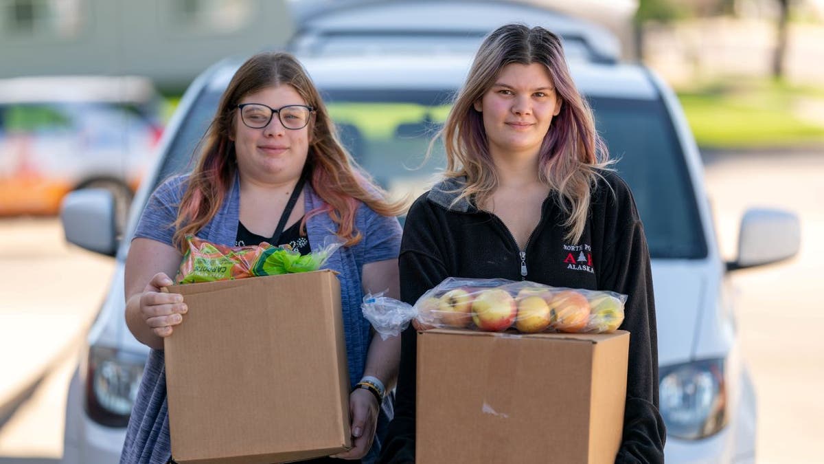 Two young women carry food in front of vehicles at a Feeding South Dakota location