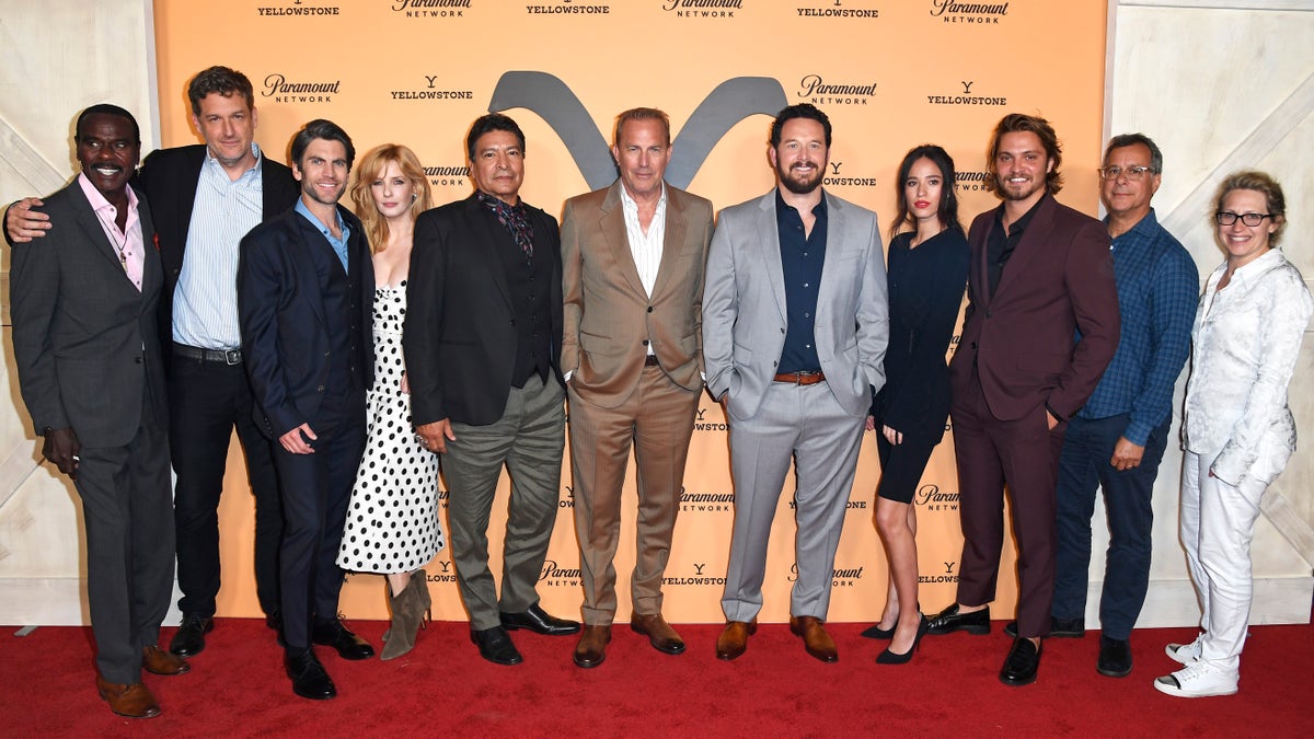 "Yellowstone" cast on the red carpet centered by Kevin Costner in a tan suit and white shirt in front of an orange "Yellowstone" backdrop