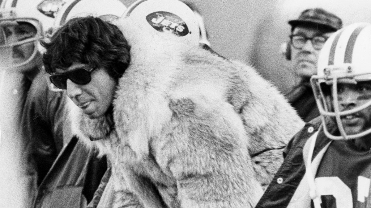 Joe Namath sits on the bench in a mink coat
