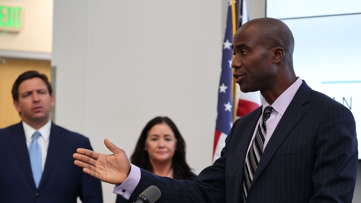 Florida Surgeon General Joe Ladapo in a pin-stripe suit with Gov. Ron DeSantis listening behind him