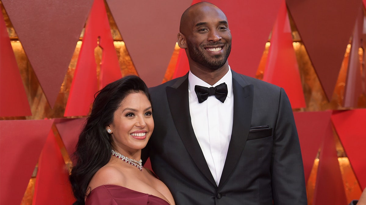 Vanessa Bryant in a red dress and Kobe Bryant a black suit in front of a red-and-gold Oscars background