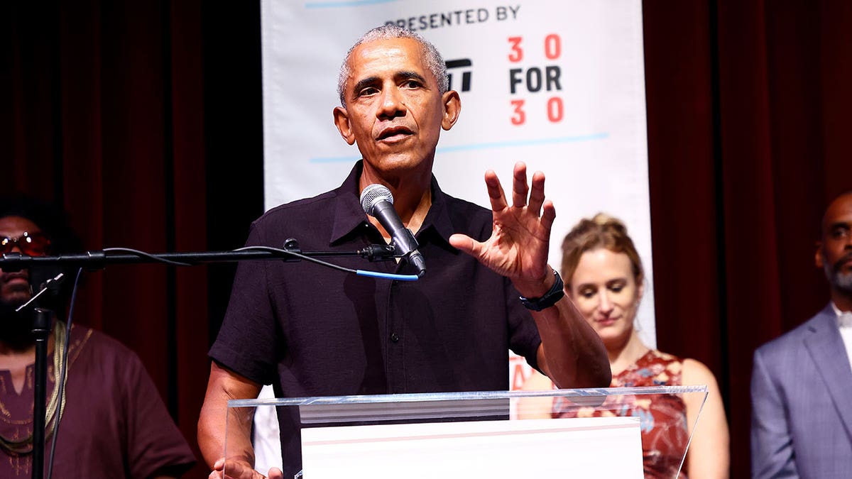 Barack Obama wearing a black polo with his hand up while speaking into a microphone at a podium