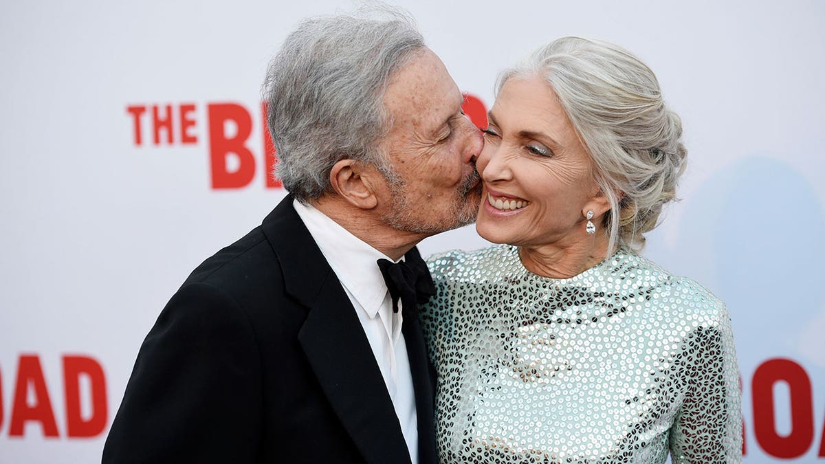 Bert Fieds kisses his wife, Barbara Guggenheim, on the cheek during a red carpet event with Barbara in a silver dress and Bert in a tuxedo