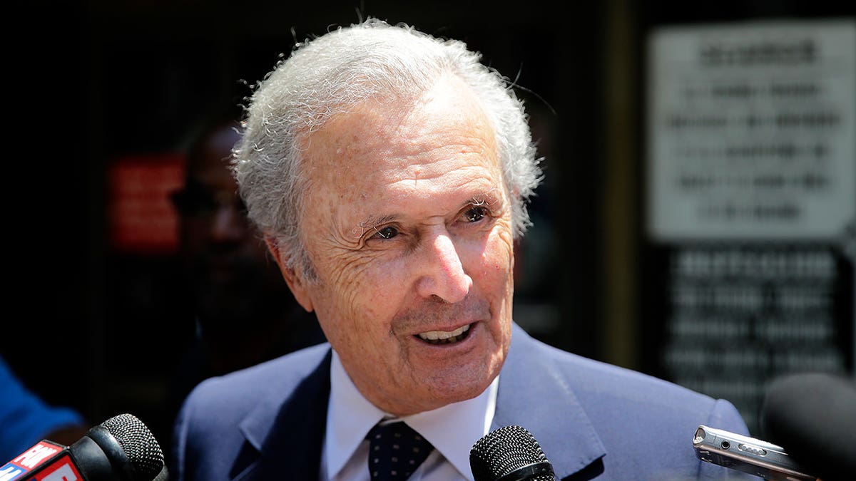 Bert Fields stands outside in daytime surrounded by microphones and reporters, dressed in a navy blue suit and tie