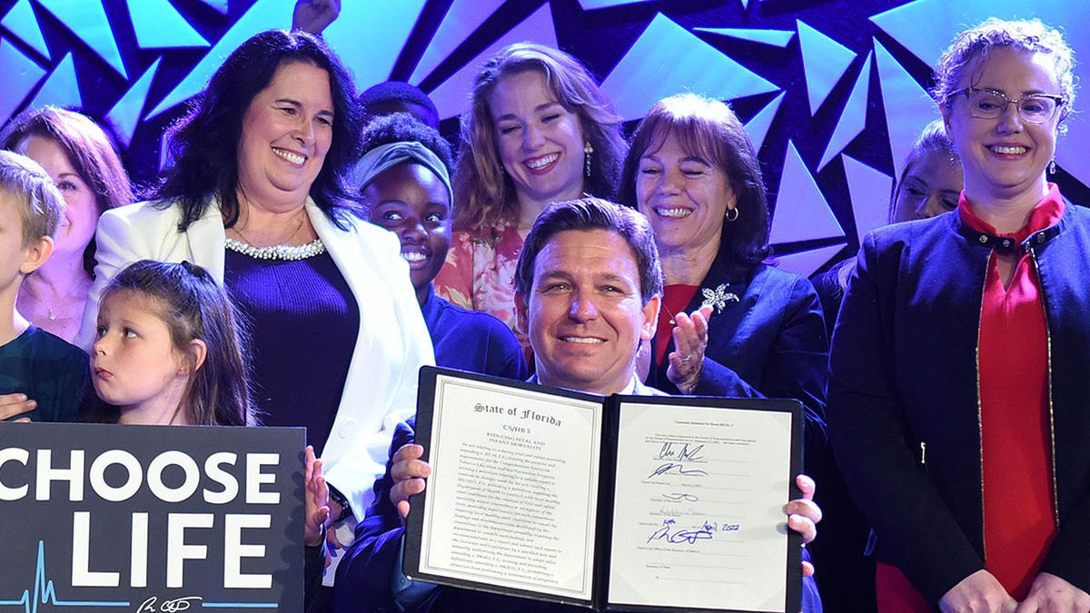 Fl Gov. Ron DeSantis holds up signed abortion ban with people around him smiling and clapping
