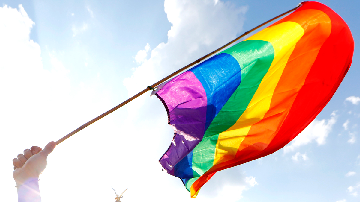 Photo shows a person holding a bright colored pride flag with blue sky and clouds in the background 