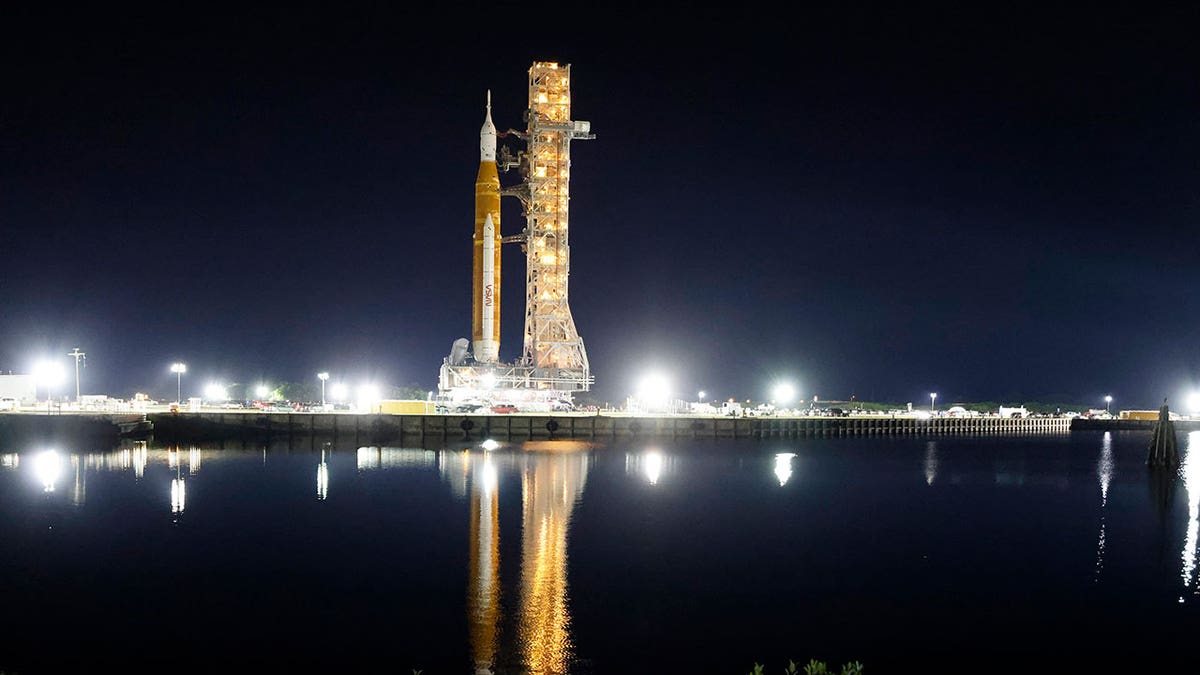 NASA's Artemis moon rocket seen on its launch pad from a distance, with dark skies overhead and its reflection in a water