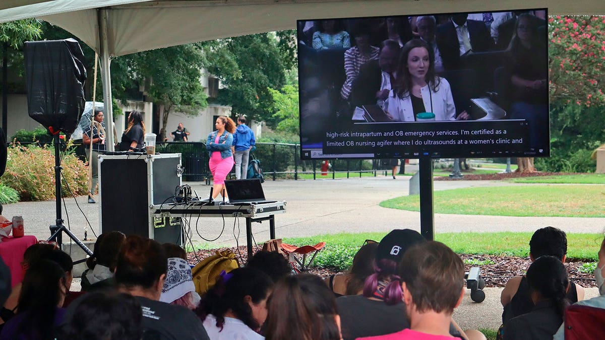People sit underneath a cover outside watching a screen with a woman speaking into microphone showing on the television