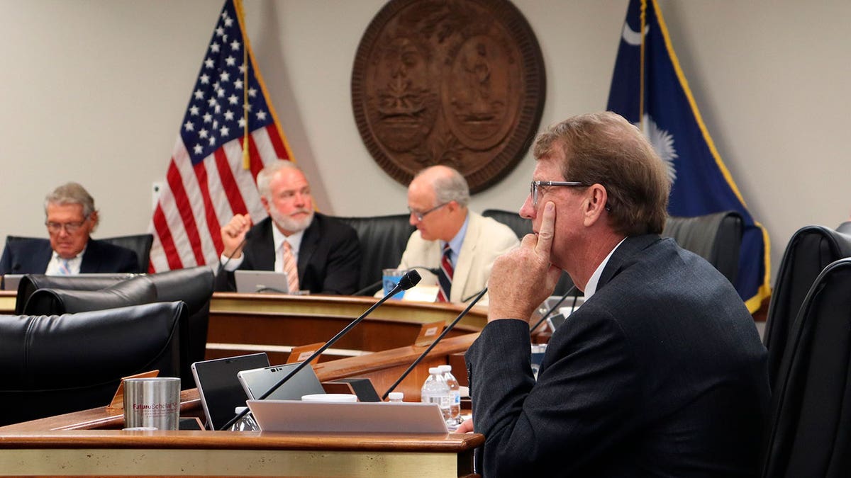 Sen. Richard Cash sits in front of a mic wearing a suit and tie with two other men sitting in the background