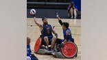 One player sitting in a wheelchair holding the ball. Another player attempts to block his pass.