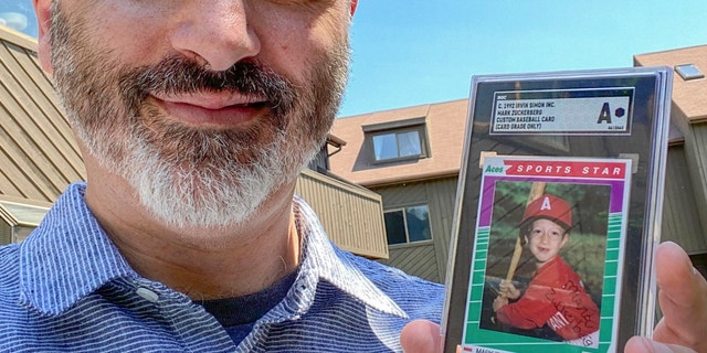 Allie Tarantino holding a baseball card featuring a very young Mark Zuckerberg grinning in a red jersey and gripping a bat. For 30 years Tarantino kept the baseball card and filed it away in his basement, not knowing Zuckerberg would someday become a household name.
