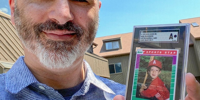Allie Tarantino holding a baseball card featuring a very young Mark Zuckerberg grinning in a red jersey and gripping a bat. For 30 years Tarantino kept the baseball card and filed it away in his basement, not knowing Zuckerberg would someday become a household name.