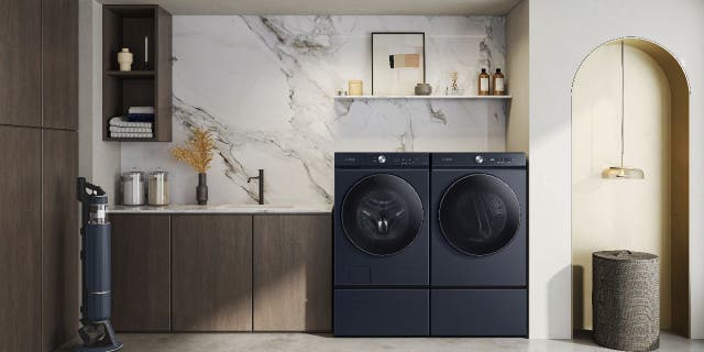 This image provided by Samsung shows a laundry room with a dark blue washer and dryer. The room is decorated with a marble-patterned wall, a stone counter, and dark wood shelves and cabinets. It is accented with a woven hamper and textured rug.