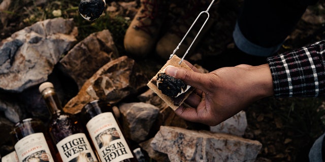 A camper makes a s'more over several High West Campfire whiskey bottles.