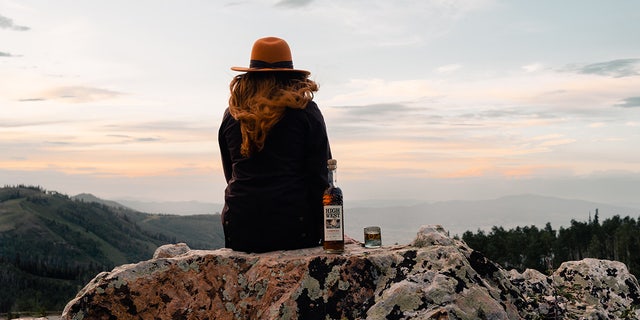 A woman sits on a mountain ledge with a bottle of High West Distillery's Campfire whiskey.