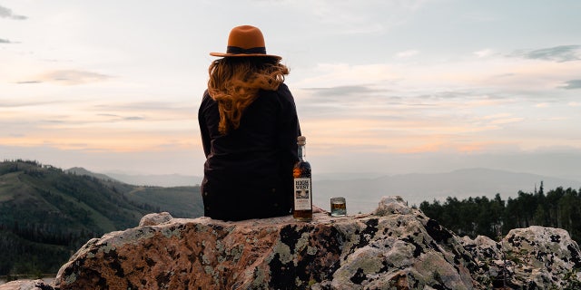 A woman sits on a mountain ledge with a bottle of High West Distillery's Campfire whiskey.