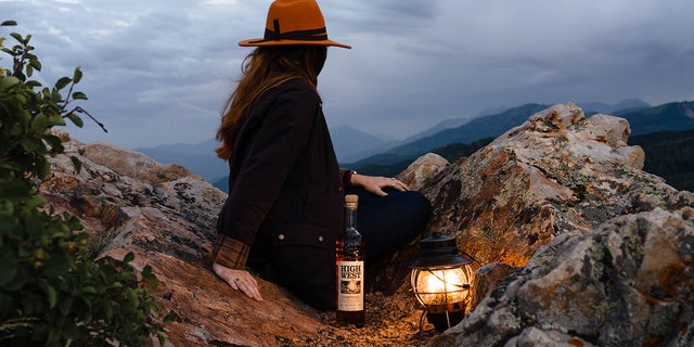 A woman takes in the mountain range view with a bottle of Campfire whiskey by High West Distillery.