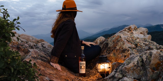 A woman admires the view of the mountain range with a bottle of Campfire Whiskey from High West Distillery.