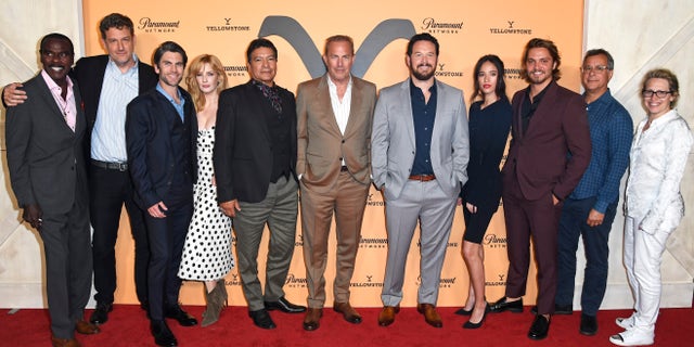 "Yellowstone" cast on the red carpet centered by Kevin Costner in a tan suit and white shirt in front of an orange "Yellowstone" backdrop