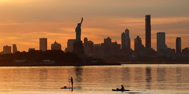 Two people paddle past the Statue of Liberty and the skyline of Brooklyn as the sun rises in New York City on August 15, 2022, as seen from Jersey City.