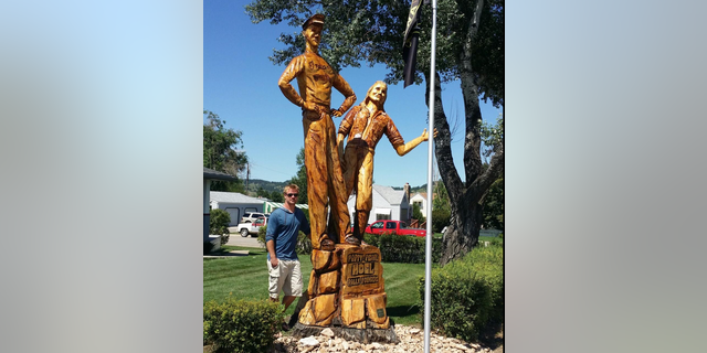 Jarrett Dahl of Dahl's Chainsaw Art poses next to his tribute to Pappy and Pearl Hoel carved from a giant silver cottonwood tree. The tree carving is outside Pappy's Vintage Cycles, once the Hoels' motorcycle shop.