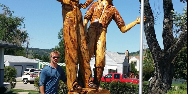 Jarrett Dahl of Dahl's Chainsaw Art poses next to his tribute to Pappy and Pearl Hoel carved from a giant silver cottonwood tree. The tree carving is outside Pappy's Vintage Cycles, once the Hoels' motorcycle shop.