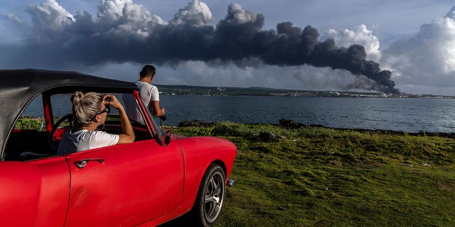 People watch a huge plume of smoke rise from the Matanzas Supertanker Base as firefighters work to douse a fire, which started during a thunderstorm the night before, in Matanzas, Cuba, Aug. 6, 2022. Cuban authorities say lightning struck a crude oil storage tank at the base, sparking a fire that ignited four explosions that injured more than 121 people, left one person dead and 17 missing. (AP Photo/Ramon Espinosa)