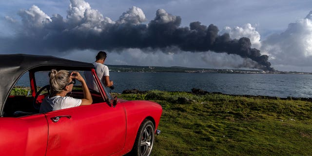 People watch a huge plume of smoke rise from the Matanzas Supertanker Base as firefighters work to douse a fire, which started during a thunderstorm the night before, in Matanzas, Cuba, Aug. 6, 2022. Cuban authorities say lightning struck a crude oil storage tank at the base, sparking a fire that ignited four explosions that injured more than 121 people, left one person dead and 17 missing. (AP Photo/Ramon Espinosa)