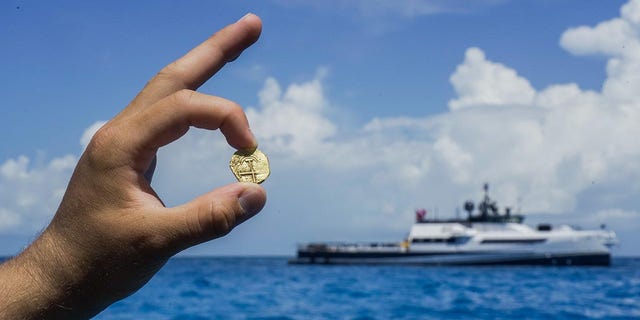 An explorer holds up a gold coin found in the Bahamas as an Allen exploration boat can be seen in the distance.