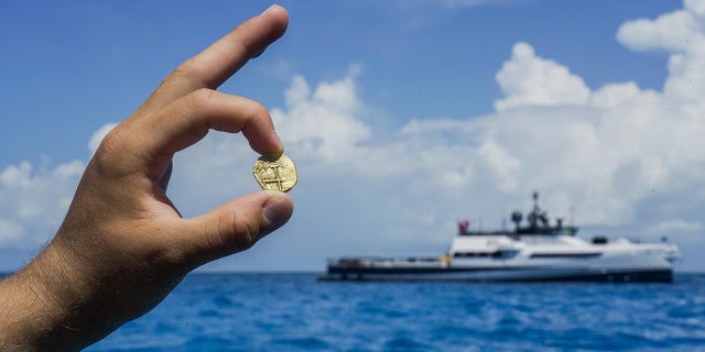 An explorer holds a gold coin found in the Bahamas as an Allen Exploration boat can be seen in the distance.