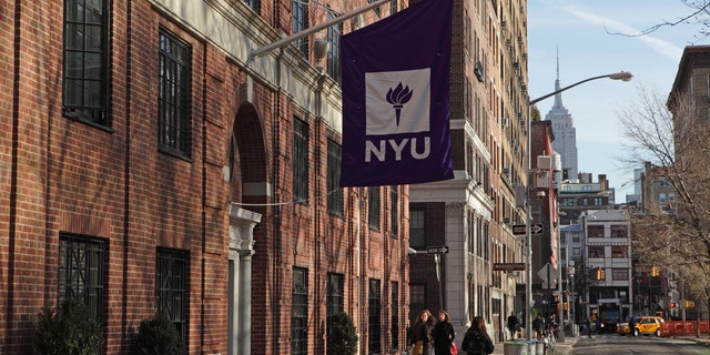 New York, NY, USA - March 22, 2016: New York University buildings on Macdougal Street at 37 Washington Square West. A purple flag with the NYU logo hangs above an entrance to one of the college buildings. In the distance, the Empire State Building appears above buildings on West 8th Street.