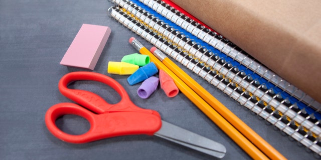 A pair of safety scissors is seen near pencils, erasers and spiral notebooks. Small children typically use safety scissors to practice cutting safely.