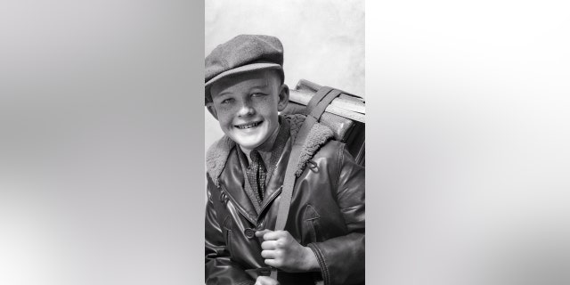 A smiling boy wears a wool newsboy cap, a leather bomber jacket and a necktie while holding a book strap.