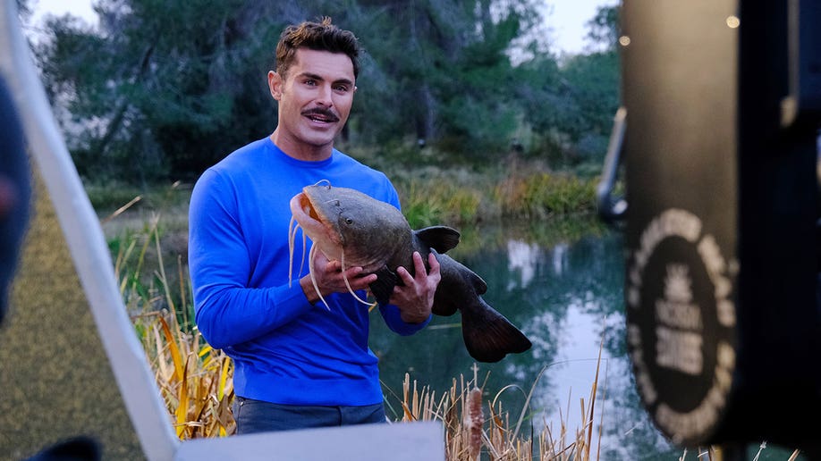 Zac Efron holding a fish while filming for "Down to Earth"