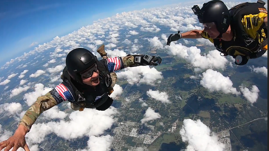 A man with a prosthetic left arm, wearing a helmet and military uniform free falling in the sky