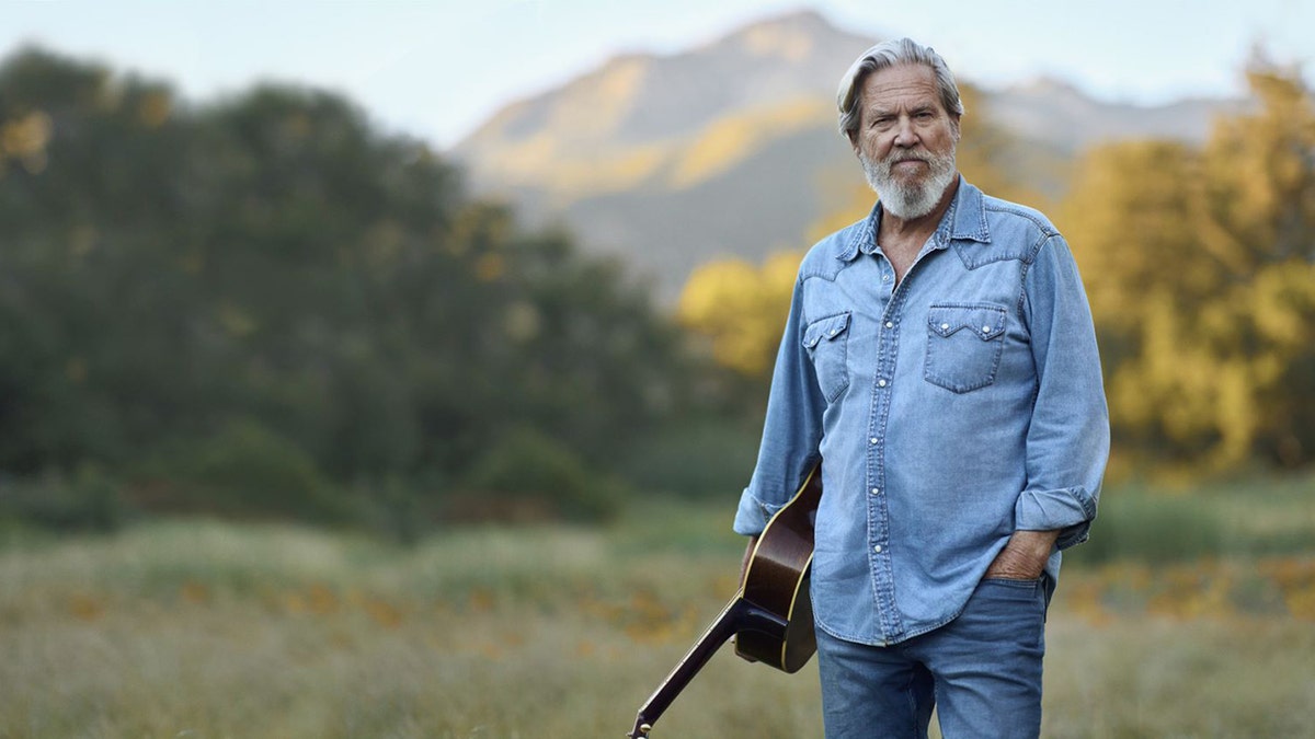 Jeff Bridges holds guitar while posing in the mountains 