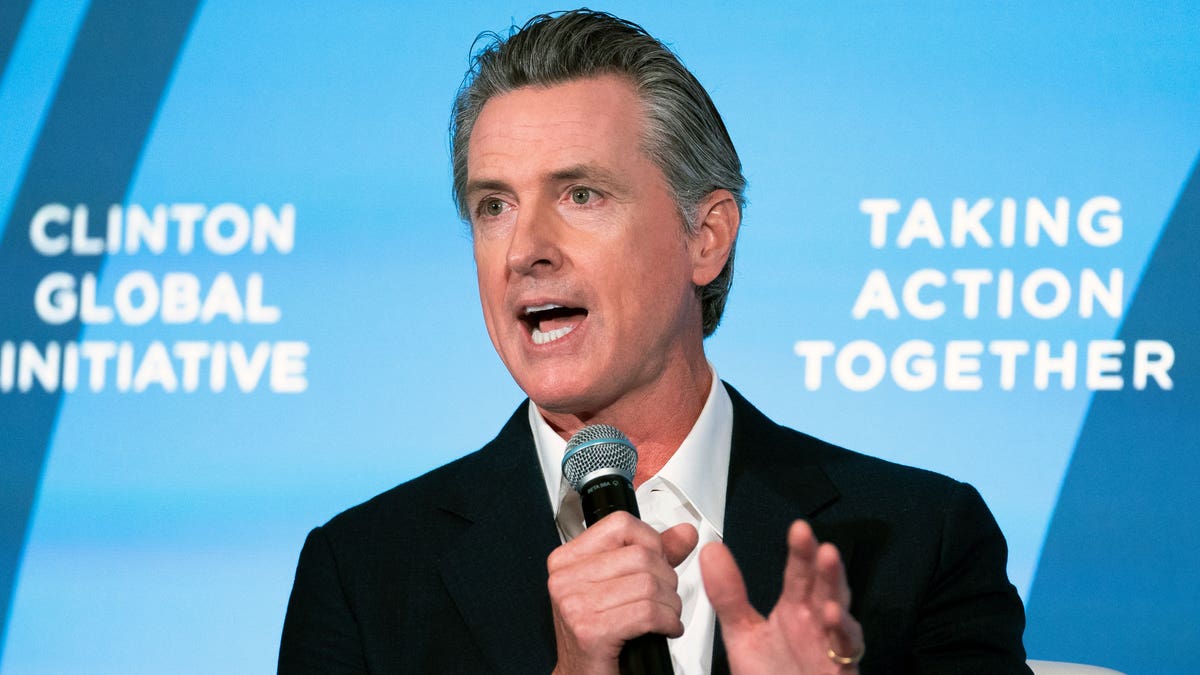 Governor Newsom holding a mic, speaking in front of a blue background