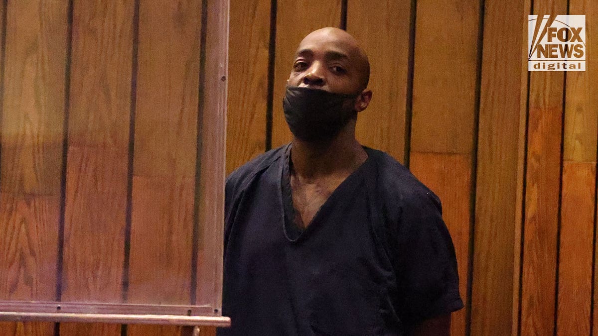 Mario Abston wears a black jumpssuit, black mask while standing in front of a wooden paneled wall