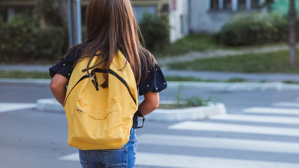 Young student with yellow book bag crosses the street.