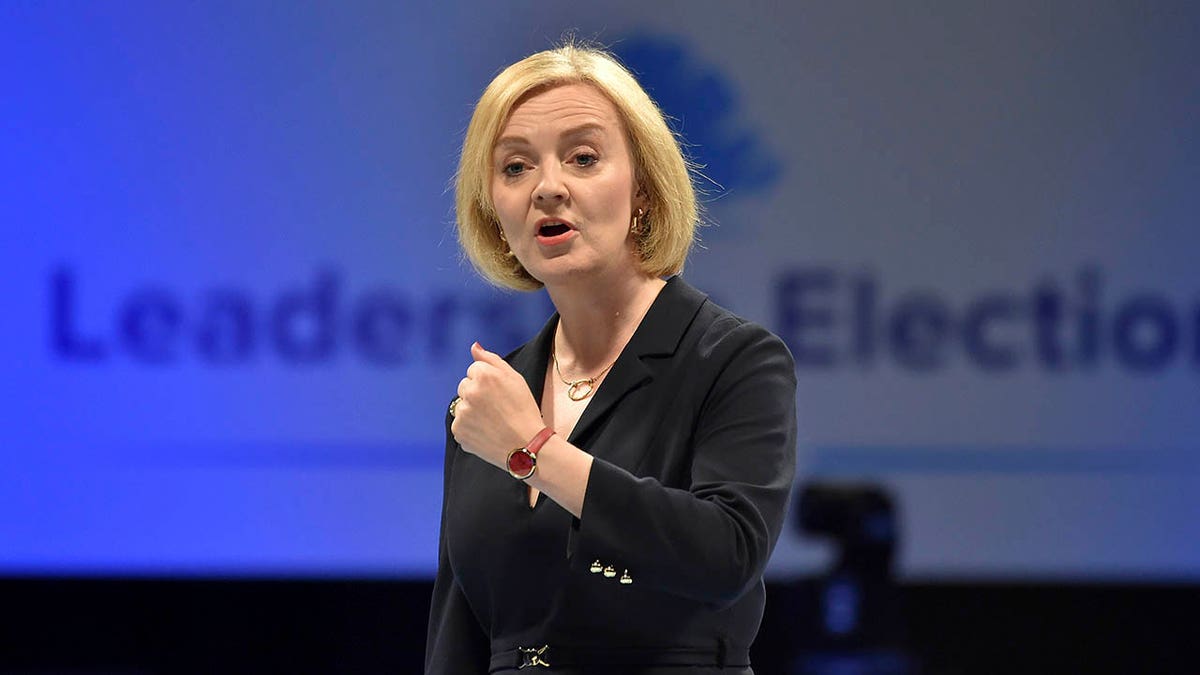 Liz Truss speaking wearing a dark suit in front of a blue/gray background