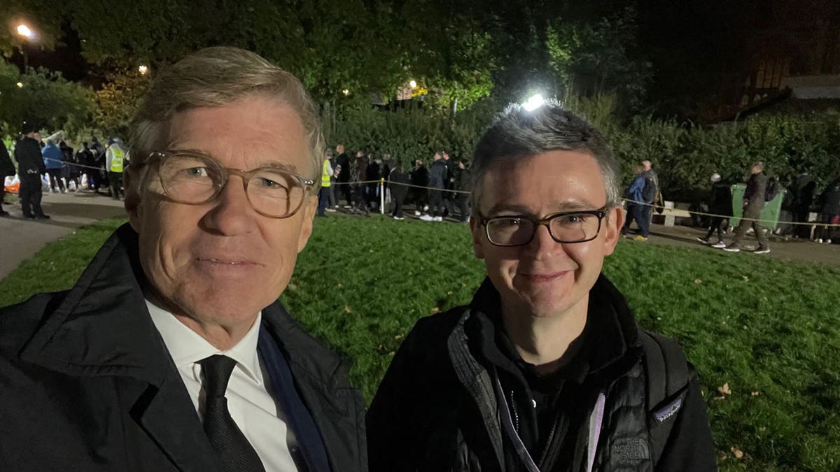 Greg Palkot wearing a suit and tie, Simon Owen wearing a jacket and backpack while outside during evening