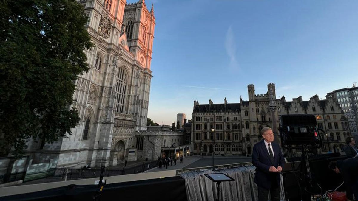 Greg Palkot stands in front of TV equipment wearing a suit tie out front of Westminster Abbey during evening.