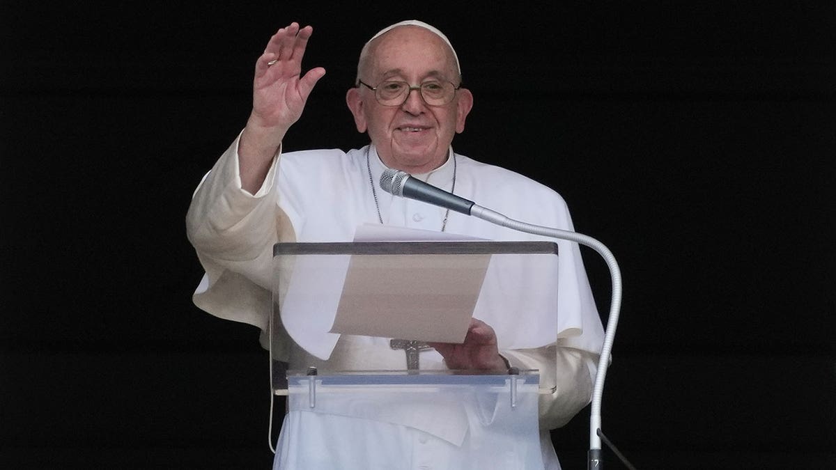 Pope Francis stands at a glass podium while holding a piece of paper and holds up his hand