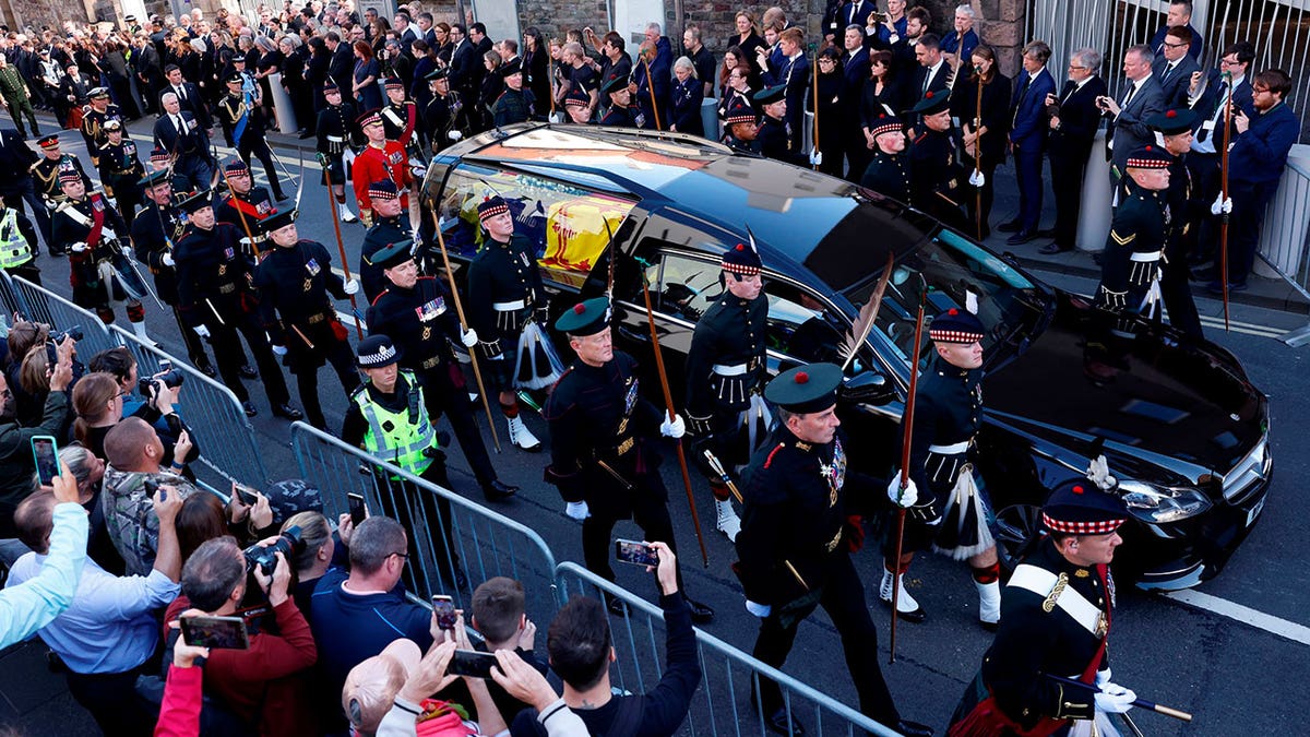 Queen Elizabeth's coffin has flowers laid on top of it as it is driven in a hearse surrounded by guards